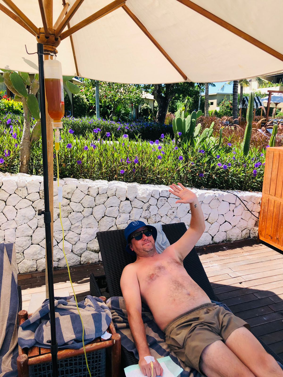 Shirtless man waving while receiving a mobile IV drip on a sun lounger in Bali.